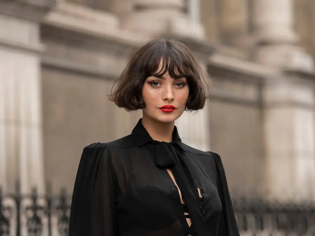 Kobieta z burzą brązowych włosów w stylu french cut, z wyrazistą szminką na ustach, w czarnej, prześwitującej bluzce.